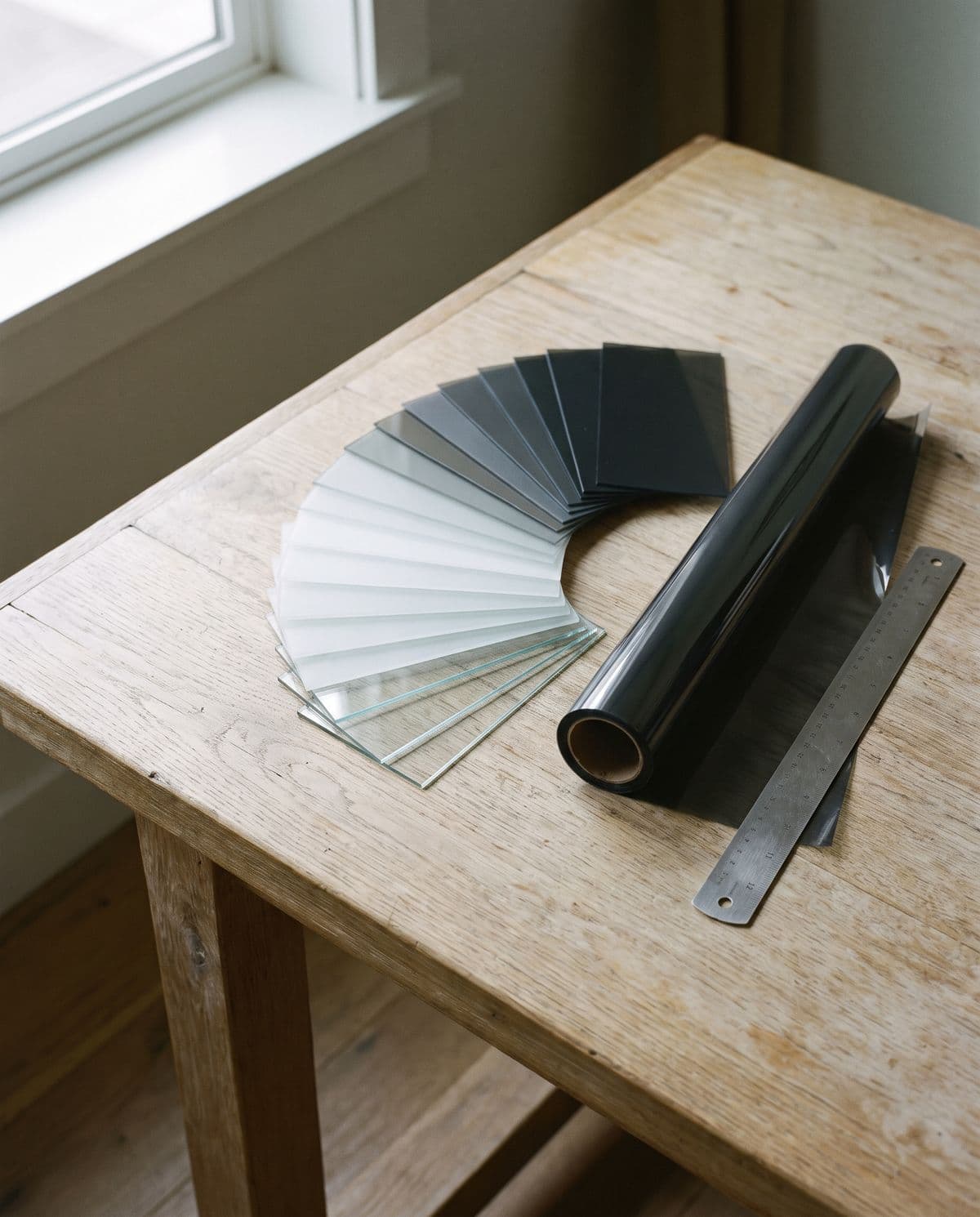 Window film sample swatches fanned across a workbench from nearly clear to deep charcoal, beside a part-unrolled core of film and a steel straight edge