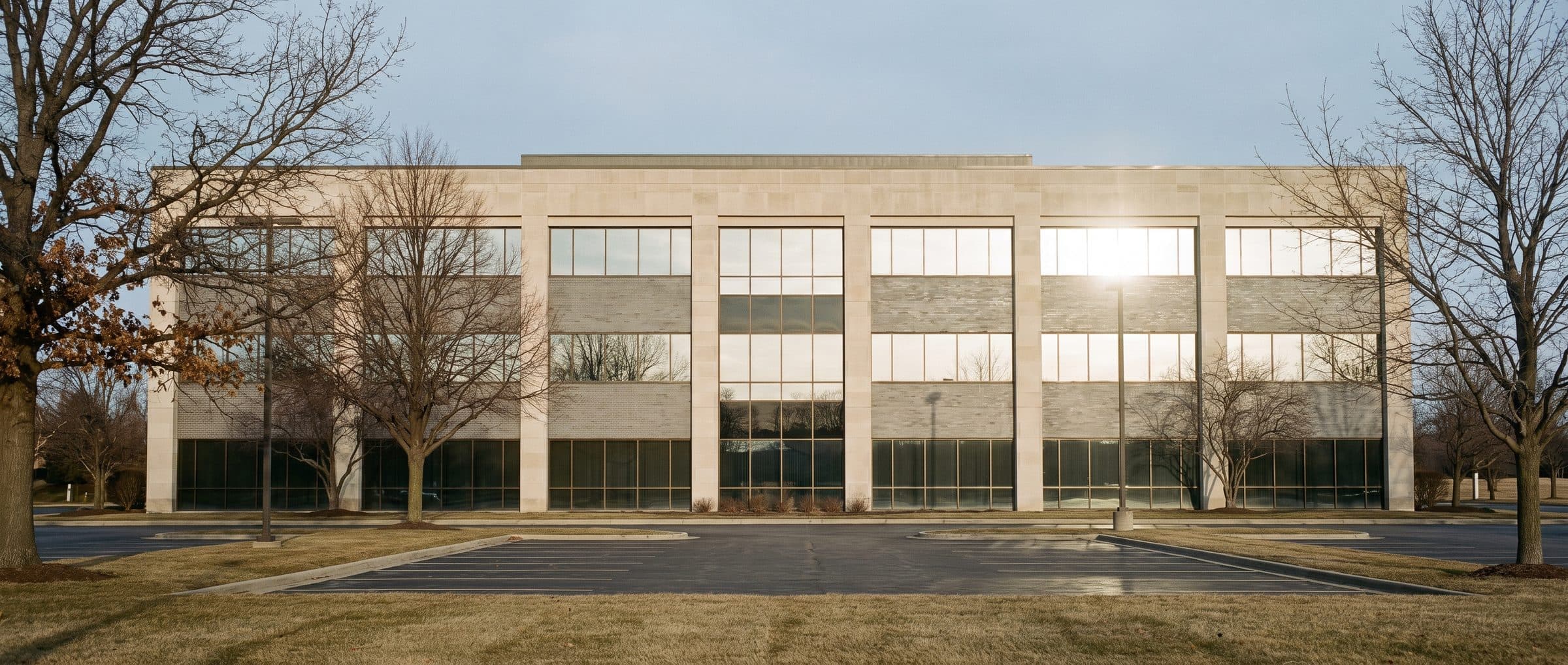 A three-storey suburban office building photographed straight on in late afternoon sun, its untreated glazing glaring white where the sun strikes it