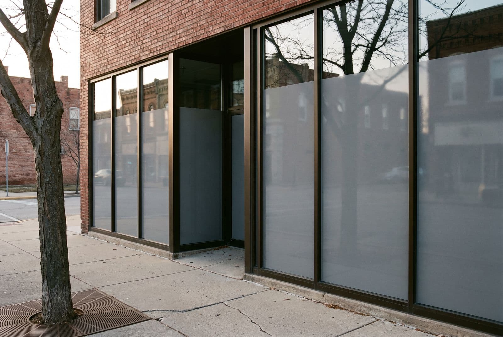 A street-level storefront whose full-height glazing now carries privacy film, reading from the sidewalk as an even calm plane that still reflects the street